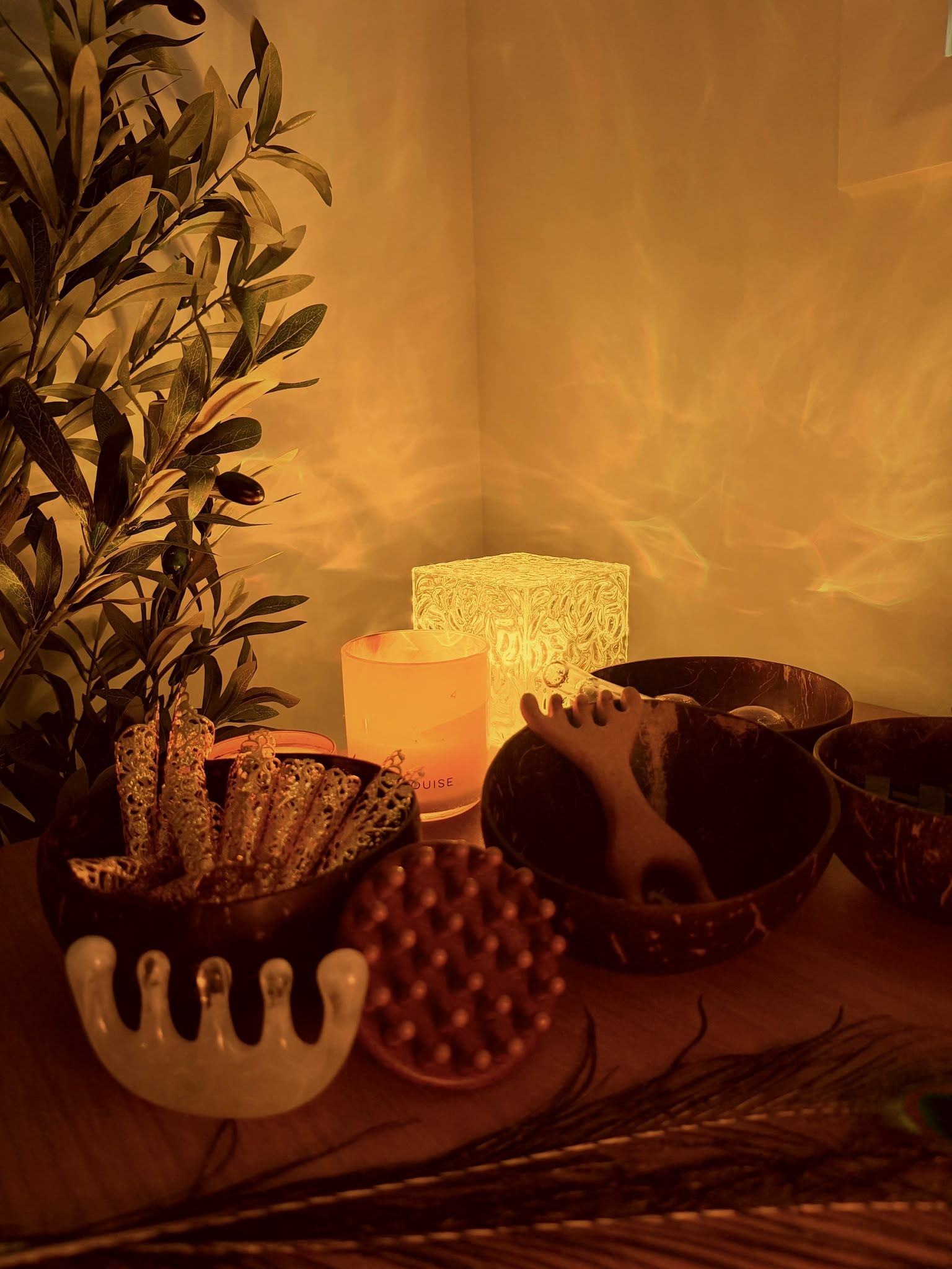 Session tools by candlelight: a peacock feather, coconut bowls, a scalp brush and comb-style massagers beside a lit candle and an olive branch.