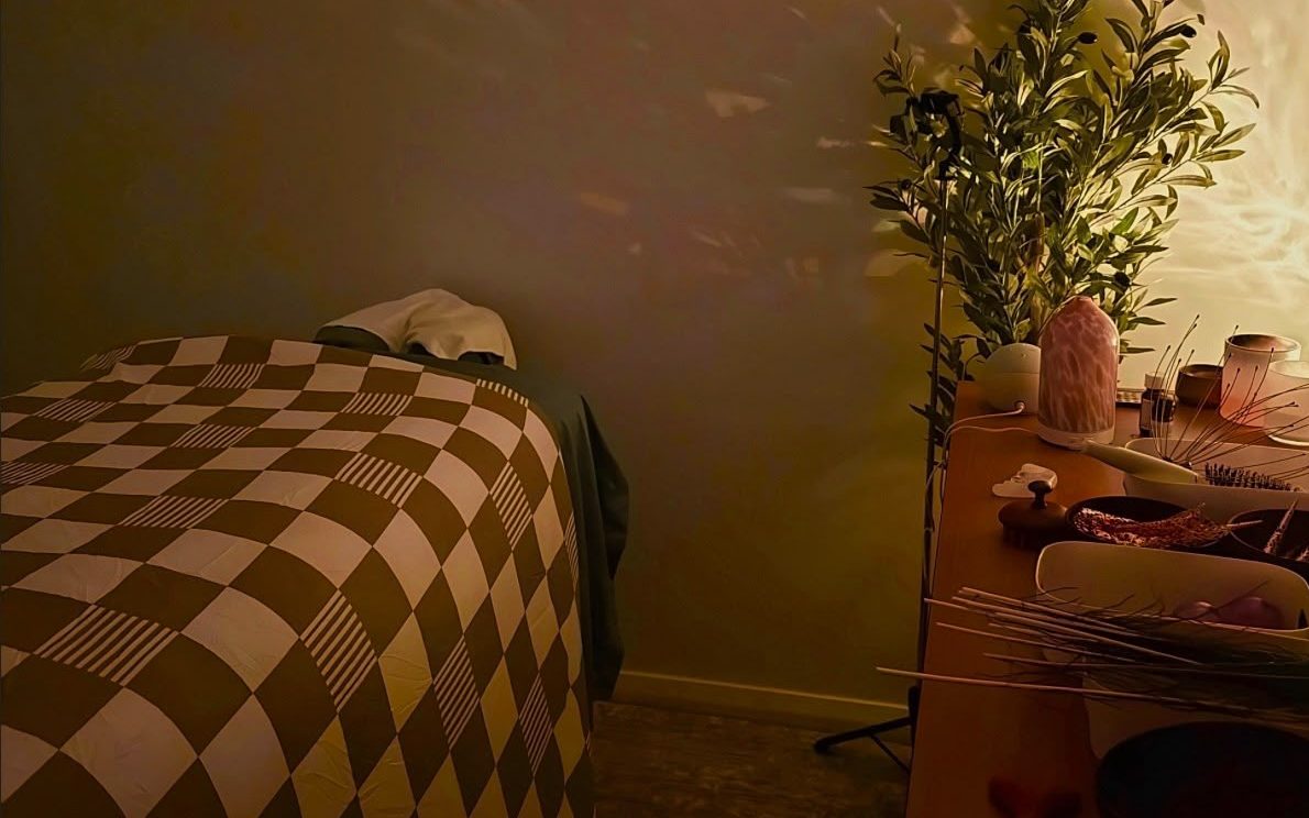 The low-lit treatment room: a bed with a checked blanket, leaf shadows on the wall, an olive tree and a table of tools.
